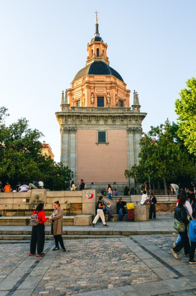 Historic architecture with a domed tower, surrounded by lively visitors and green trees in a vibrant Madrid plaza.