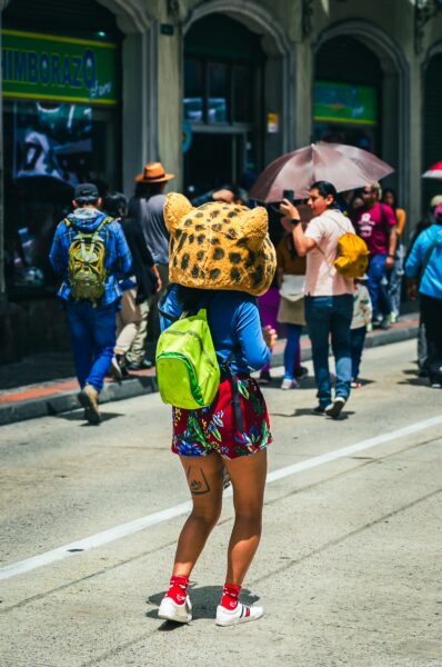 Colorful parade scene in Quito, Ecuador, featuring a person with a leopard print headpiece and a bright green backpack walking among crowds.