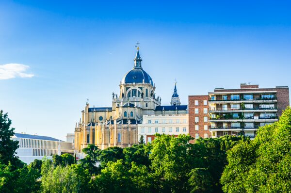 Historic architecture with a grand dome rises amidst green trees under a clear blue sky in Madrid, Spain.