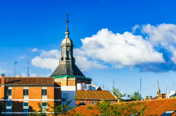 Historic rooftops and a striking bell tower under a vibrant blue sky in Madrid, Spain, with fluffy white clouds drifting by.