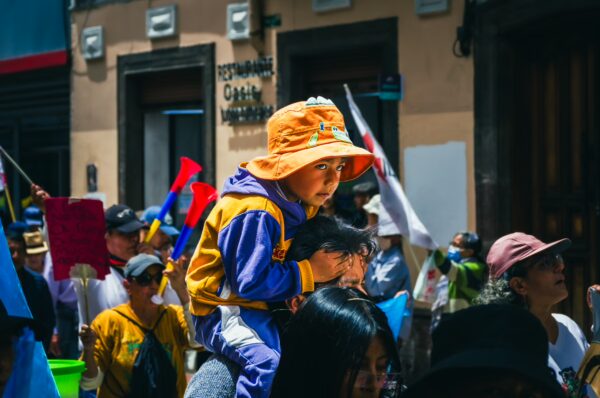 A child wearing an orange hat sits on a parent's shoulders, surrounded by a vibrant crowd with flags at the International Workers' Day parade in Quito.