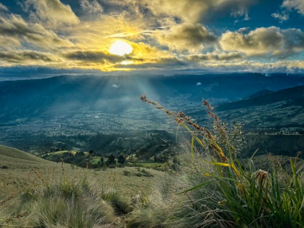 Golden rays of sunlight illuminate Imbabura Volcano, with lush green valleys and tall grasses in the foreground, Ecuador.