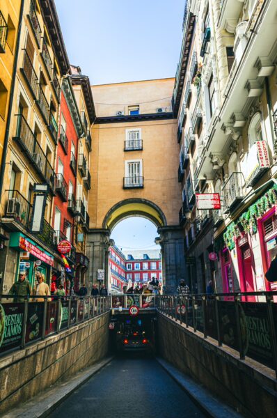Vibrant buildings frame the archway leading to Plaza Mayor, with shops lining the streets under a clear blue sky in Madrid, Spain.