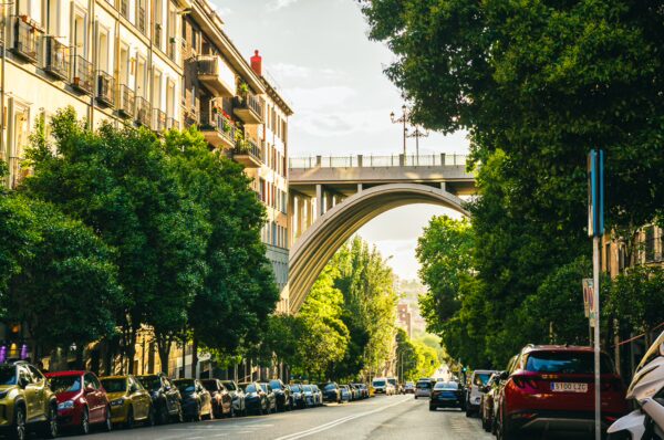 Vibrant street lined with trees and parked cars, showcasing the modern arch bridge in Madrid, Spain. Bright afternoon light enhances the scene.
