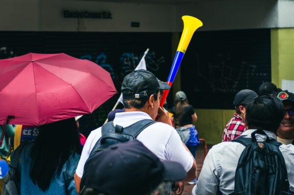 Colorful crowd celebrating International Workers' Day in Quito, Ecuador, with umbrellas and festive horns visible.