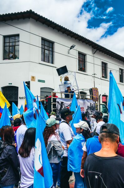 Bright blue flags wave among a crowd at the International Workers' Day parade in Quito, Ecuador, under a partly cloudy sky.