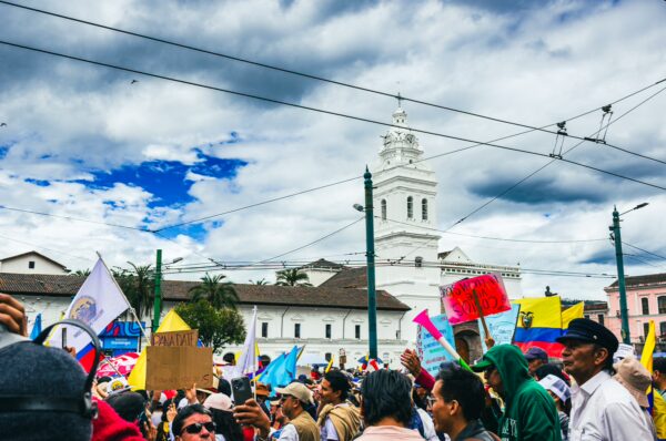 Colorful banners wave above a lively crowd during the International Workers' Day parade in Quito, Ecuador, with a historic building in the background.