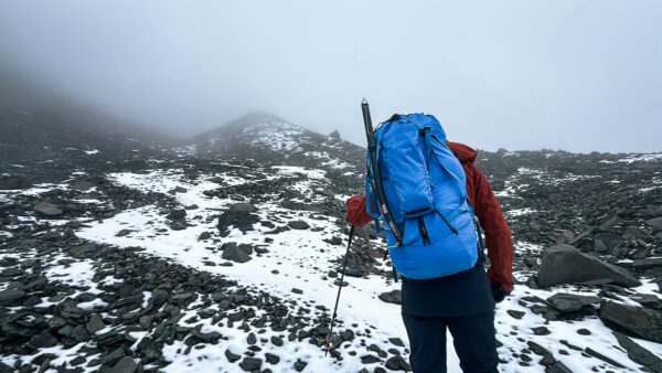 Hiker with a blue backpack navigating snowy rocky terrain on Chimborazo Volcano, Ecuador, shrouded in mist.