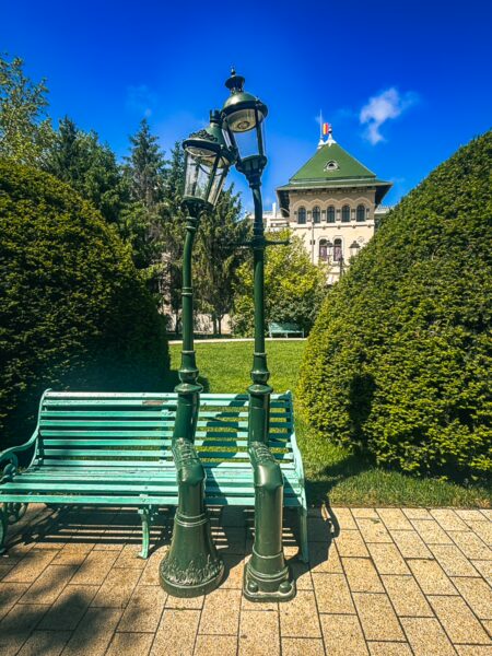Vintage lampposts with a green bench set against neatly trimmed hedges in English Park, Craiova, Romania. Bright blue sky above.