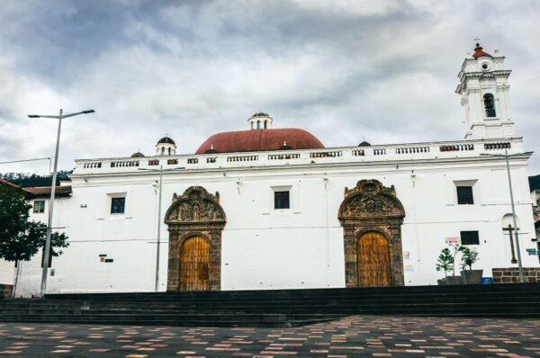 Santa Clara Monastery showcases striking white walls and ornate wooden doors under a cloudy sky in Quito, Ecuador.