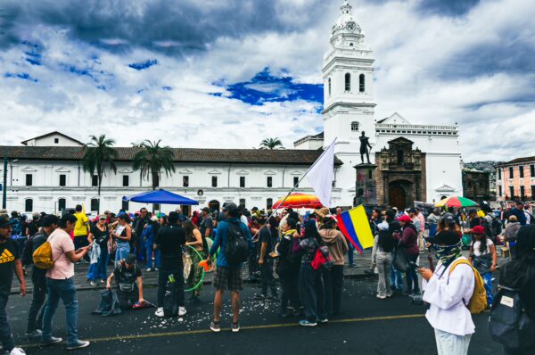 Crowds gather with flags during the International Workers' Day parade in Quito, Ecuador, with a church and cloudy skies in the background.