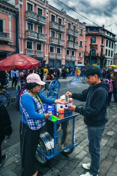 Colorful street vendor serving drinks during the International Workers' Day parade in Quito, Ecuador, with lively crowds in the background.