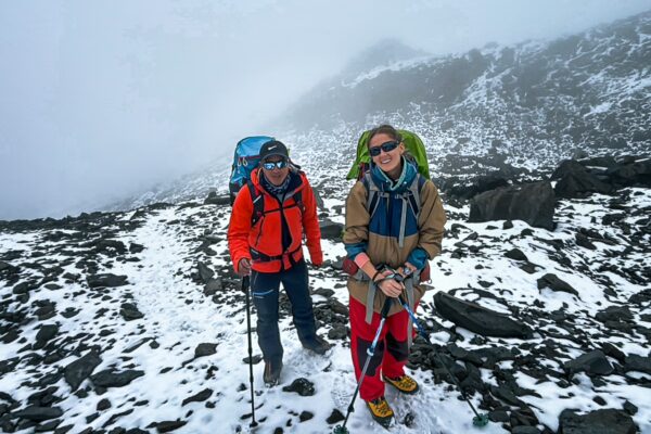 Two hikers navigate snowy terrain near Chimborazo Volcano, Ecuador, wearing colorful gear under a misty sky.