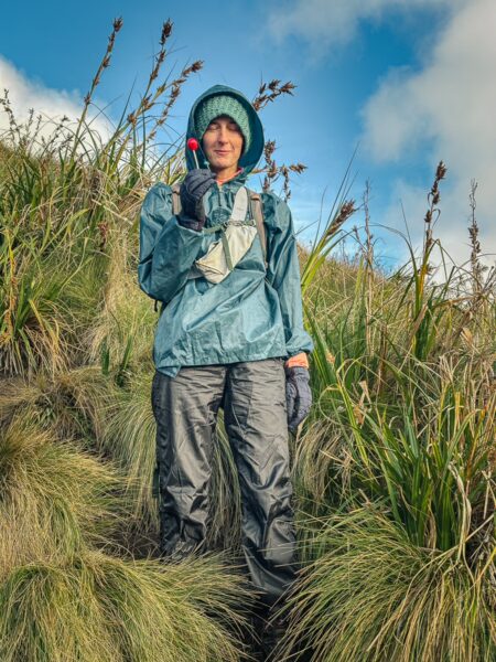 Hiker in a green jacket and gloves stands among tall grass with hints of blue sky, set against Imbabura Volcano, Ecuador.