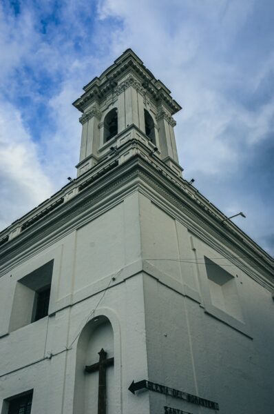 Historic white church tower with a striking bell and cross, set against a cloudy sky in Quito, Ecuador.