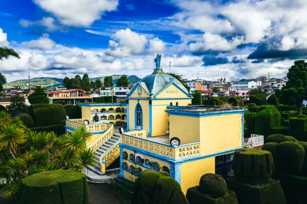 Vibrant blue and yellow chapel surrounded by lush greenery at Tulcán Cemetery in Ecuador under a dramatic sky.