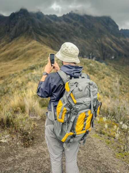 A hiker in a gray and yellow backpack stands on a trail, capturing the rugged mountains of Ruminahui, Ecuador.
