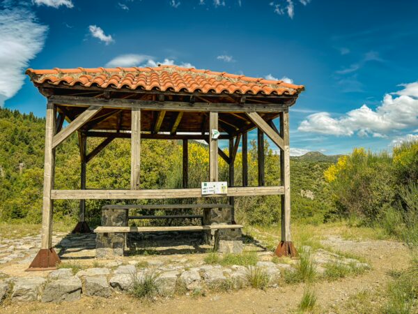 Wooden gazebo with a red tile roof set against a backdrop of lush greenery and blue skies in Vytina, Arcadia, Greece.