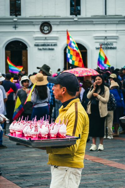 Colorful parade in Quito for International Workers' Day, featuring a vendor with pink treats and rainbow flags amidst a festive crowd.