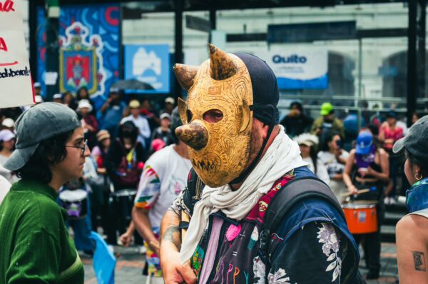 A masked participant in a colorful outfit stands in a lively crowd during the International Workers' Day parade in Quito, Ecuador.