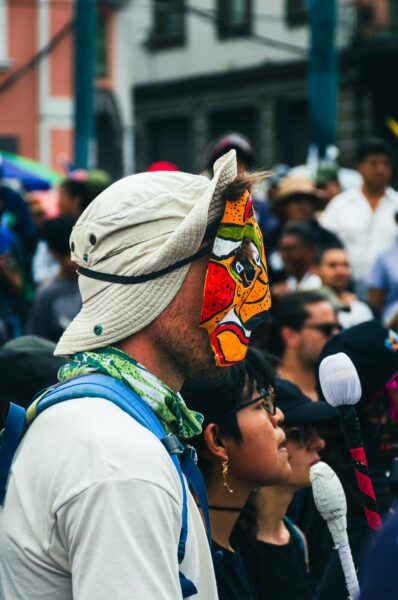 Colorful mask on a participant wearing a hat, surrounded by a diverse crowd during the International Workers' Day parade in Quito, Ecuador.