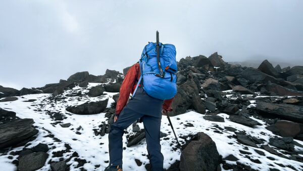 Hiker ascending rocky terrain with snow near Chimborazo Volcano, Ecuador, under a cloudy sky. Bright blue backpack stands out.