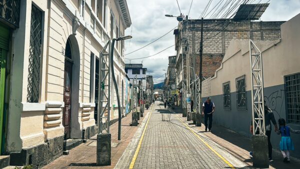 Cobblestone street lined with colonial buildings and power lines in Machachi, Ecuador, under a cloudy sky.