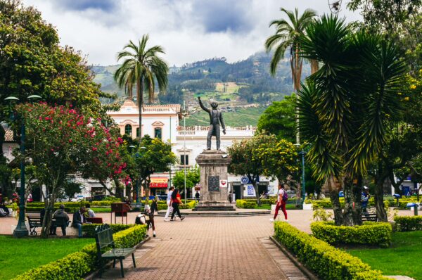 Vibrant greenery and palm trees surround a statue in Parque Pedro Moncayo, Ibarra, Ecuador, with mountains in the background.