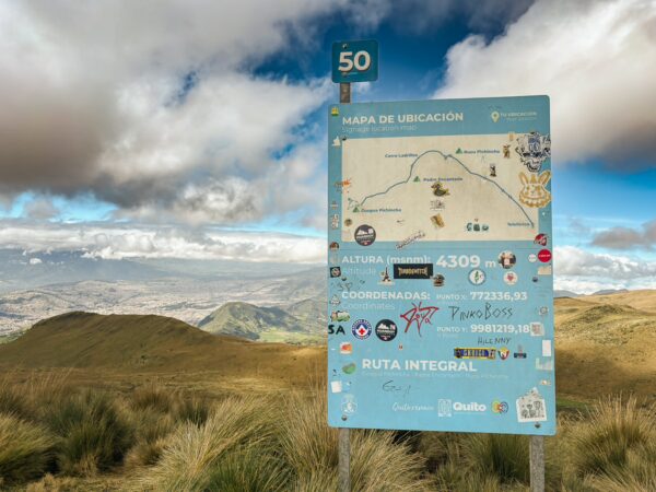 Signpost marking the route at Pichincha Volcano in Ecuador, featuring a map and altitude information against a cloudy sky.
