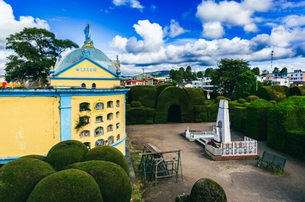 Colorful mausoleums and lush green shrubs create a serene landscape at Tulcán Cemetery in Ecuador, under a bright blue sky.