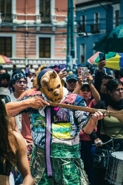 Vibrant International Workers' Day parade in Quito, Ecuador, featuring a performer in a bear mask playing a string instrument among colorful crowds.
