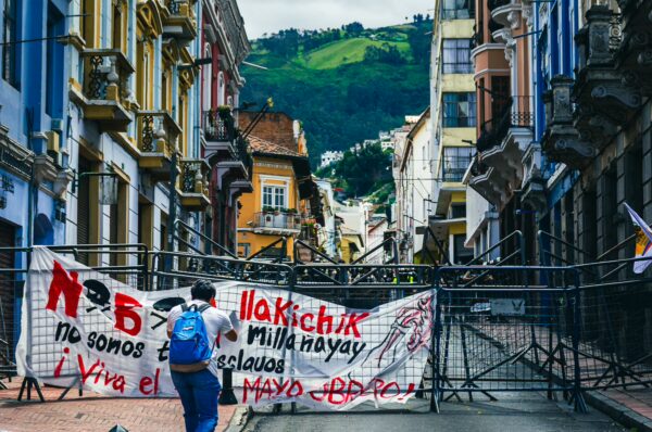 Colorful buildings line the street in Quito, Ecuador, as demonstrators pass a barricade adorned with protest banners.