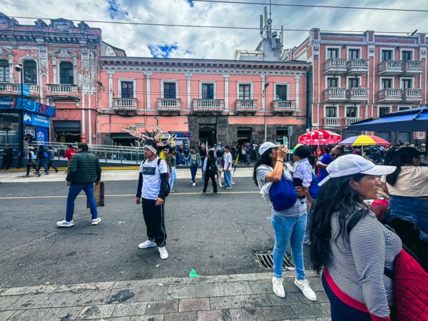 Crowds gather in colorful attire during the International Workers' Day parade in Quito, Ecuador, with historic buildings in the background.