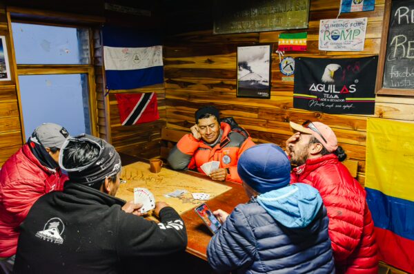 Travelers relax inside the Jose Rivas Refuge, Mount Cotopaxi, Ecuador, playing cards against a backdrop of flags and wooden walls.