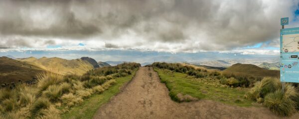 Scenic view from Pichincha Volcano features a winding dirt path, lush green slopes, and dramatic clouds over Ecuador's landscape.
