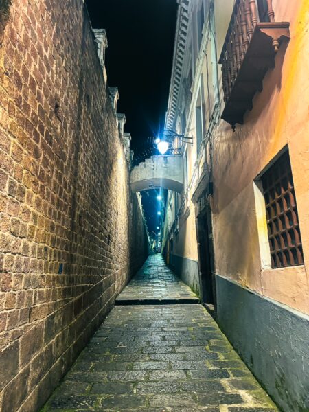 Narrow cobblestone alley lined with historical brick walls, illuminated by street lamps in La Ronda, Quito, Ecuador.