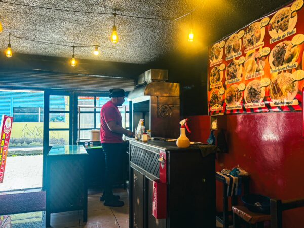 Asadero El Rinconcito features a cook preparing food in a cozy, warm-lit interior with a menu visible on the wall in Machachi, Ecuador.