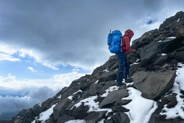 Hiker in a red jacket and blue backpack climbs rocky terrain with patches of snow on Chimborazo Volcano, Ecuador.