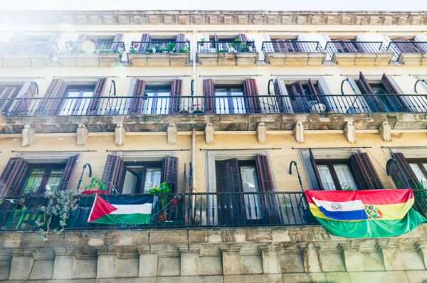 Balconies adorned with vibrant flags and lush greenery against a classic Madrid building. Bright sunlight enhances the colors.