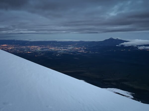 Snow-covered slopes descend towards a dimly lit valley, with distant city lights visible beneath the cloudy sky near Mount Cotopaxi, Ecuador.