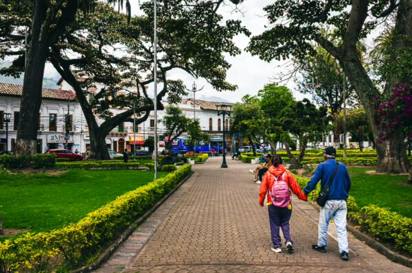 Couple strolling hand-in-hand through Parque Pedro Moncayo, showcasing vibrant greenery and quaint colonial architecture in Ibarra, Ecuador.
