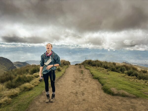 Hiker stands on a rugged path amidst green hills with dramatic clouds over Pichincha Volcano, Ecuador.