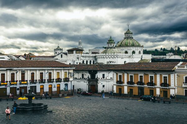 Historic architecture and a central fountain in San Francisco Square, Quito, Ecuador, set against a dramatic cloudy sky.