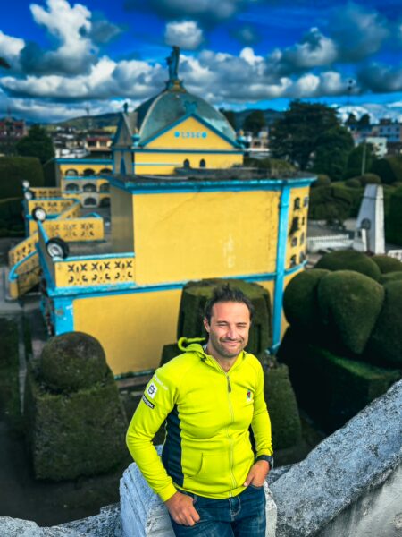 Bright yellow mausoleum stands amidst neatly trimmed shrubs at Tulcán Cemetery in Ecuador, with a vibrant blue sky overhead.