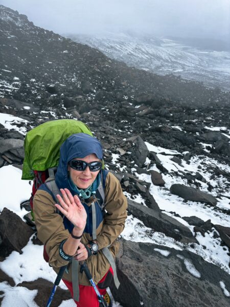 Hiker on rocky terrain with snow near Chimborazo Volcano, Ecuador, wearing sunglasses and a colorful backpack amidst a misty landscape.
