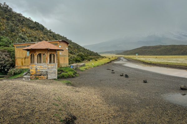 Rustic building set against a dramatic landscape of Cotopaxi National Park, featuring green hills and cloudy skies.