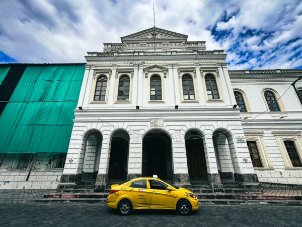 Yellow taxi parked in front of a white colonial building with intricate architecture in Ibarra, Ecuador, under a cloudy sky.