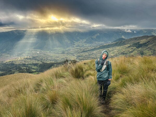 Hiker on a lush trail with sun rays breaking through clouds, overlooking the Imbabura Volcano in Ecuador.