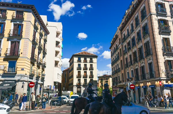 Vibrant street scene in Madrid, Spain, featuring historic buildings, blue skies, and a mounted police officer amid bustling pedestrians.