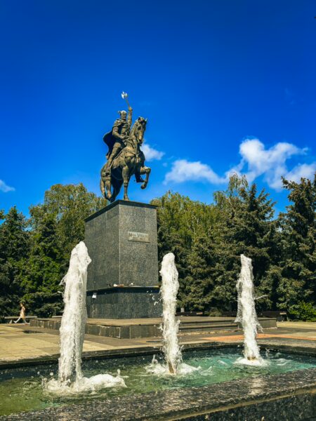 Michael the Brave statue stands proudly on a pedestal, surrounded by three vibrant fountains against a clear blue sky in Craiova, Romania.